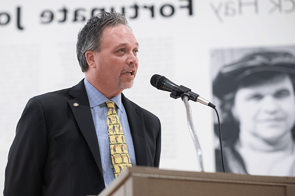 Provost Olsen speaking at a podium with a microphone, dressed in a suit and patterned tie, against a backdrop featuring large text and a black-and-white photograph. The setting appears to be a formal or commemorative event